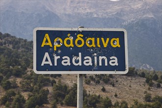 Damaged road sign with Greek and Latin lettering in front of a mountain landscape, Aradena Gorge,