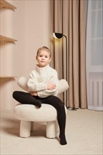 A little girl in warm sweater sits on a soft chair with one leg crossed and her arms crossed over