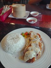 A plate of rice with chicken cordon bleu and mixed vegetables topped with creamy sauce