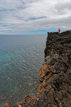 A brave person stands on the edge of a high volcanic cliff above the sea, lava rocks coastal hiking