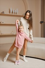 A joyful mother and her young daughter dancing together in a cozy living room. The daughter wears a