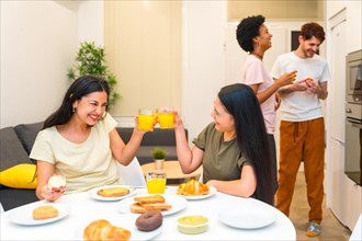 Two women toasting with juice while having breakfast with friends at home