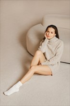 Young woman with long bare legs in socks wearing knitted sweater sitting on the floor near a sofa