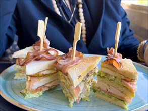 Woman Sitting Around a Table in Restaurant and Eating Club Sandwich with Bacon in a Sunny Day in