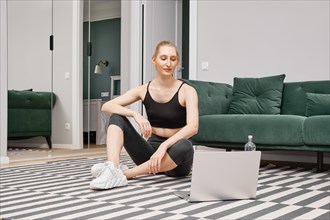Young woman sits on the floor of her apartment and watching video record with sport exercises