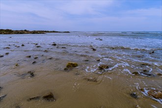 Landscape of ocean shoreline with waves washing up on rocks in the sand and a small rocky outcrop