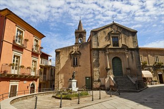 A small square with an old church and statues under a blue sky in a town, Novara di Sicilia,