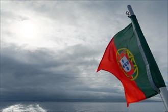 Portuguese flag waving majestically against a cloudy sky over the sea, Horta, Faial Island, Azores,