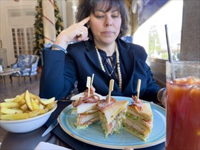 Elegant Woman Sitting Around a Table in Restaurant and Eating Club Sandwich with Bacon and French