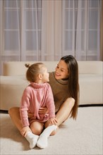 A young mother and daughter sit on the floor in a cozy living room, with a couch and curtains in