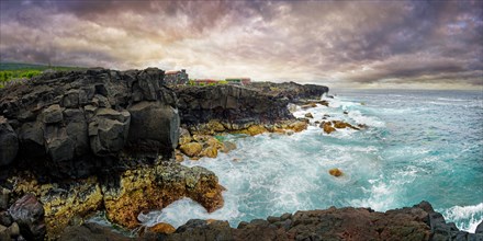View of the wild coastline with rugged cliffs of lava rock and roaring waves under a grey sky,