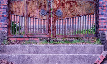 View of stairs leading to a chained rusted gate with red brick columns on each side located at old