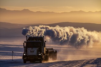Truck on snowy track with large exhaust cloud in front of mountains in winter, evening light,