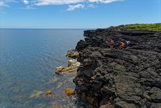 Three people hiking on rocky coastline next to the clear blue sea water, lava rocks coastal hiking