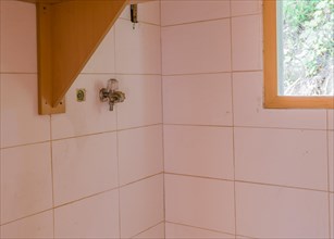 Water faucet on white tiled wall next to wooden shelf and window in laundry room of abandoned house