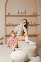 Happy young mother in warm sweater and socks sitting on top of a white ottoman next to her daughter