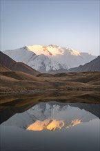 White glaciated and snowy mountain peak Pik Lenin at sunset, mountains reflected in a lake between