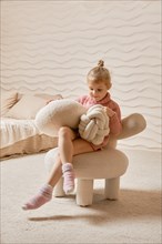 Little girl playing with stuffed pillows in her room, sitting on soft chair and knocking pillows