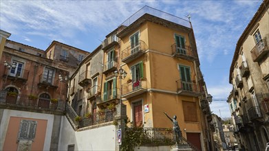 A street corner in a town with a bronze statue and traditional balconies, Novara di Sicilia,