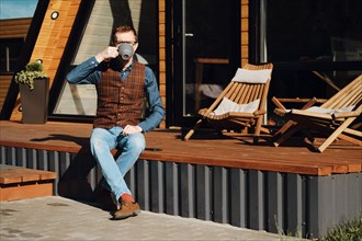 Middle-aged man sitting on terrace of wooden cabin and drinking tea in the morning