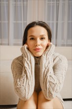 A contemplative woman, wrapped in a cozy knitted sweater, sits next to bed. Her hands wrap around