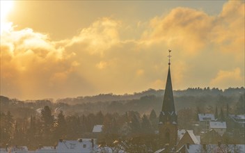 The sun sets behind a wintry forest with a church tower, radiating mild light, Wuppertal Vohwinkel,