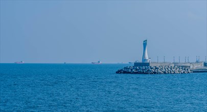 White lighthouse on concrete pier under slate gray sky with cargo ships in background