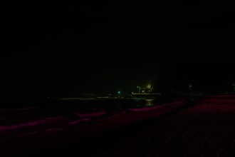 Night scene of deserted beach in darkness with waves coming into shore and street lights over road