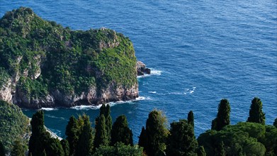 Cliff on a coast with lush greenery surrounded by the blue sea with visible waves, Taormina,