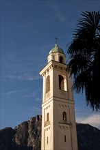 Church Bell Tower of San Zenone Against Blue Sky and Mountain Peak San Salvatore and Palm Leaf in