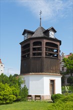 A wooden clock tower stands under a blue sky surrounded by trees and grass, Clock Tower, Oratorony,