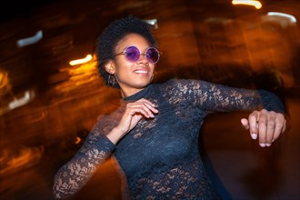 Night photo with flash and motion of an african woman with sunglasses dancing in the city street at