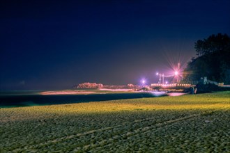 Night scene at beach bathed in soft light with unrecognizable people and pier lit up by street