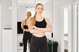 Young woman in sportswear stands in living room with her arms crossed on her chest