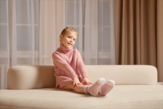 Cute little girl in pink sweater sitting on the sofa with her arms and legs stretched forward