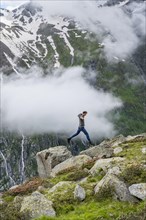 Mountaineer jumping from one rock to the next, cloudy mountain landscape with blooming alpine