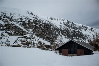 A solitary cabin sits beneath a rugged, snowy mountain landscape under an overcast sky. Poland,
