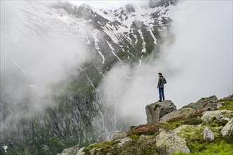Mountaineer standing on a rock, cloudy mountain landscape with blooming alpine roses, view of rocky