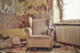 A worn armchair in front of a crumbling wall with falling wallpaper in an abandoned room,
