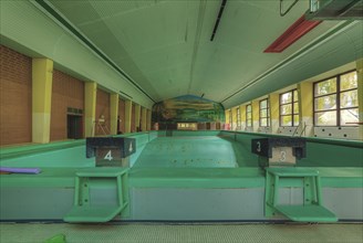 An abandoned indoor swimming pool with starting blocks and a mural in the background, Bad am Park,