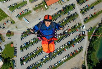 Paragliding pilot with orange and blue clothes, flying high over parking lot of Brauneck cable car,