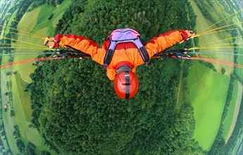 Paraglider pilot with orange clothes and red helmet flying in 500 ft above a forest and meadows,