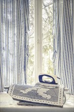 An old iron on a lace fabric in front of a window with blue curtains, urologist's villa Dr Anna L.,
