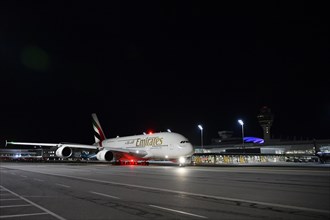 Emirates Airlines Airbus A38-800 taxiing on apron in front of Terminal 1 with tower at night,