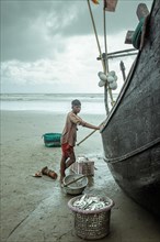 Fisherman by his boat on the beach with the catch of the day during a monsoon shower, Cox's Bazar,