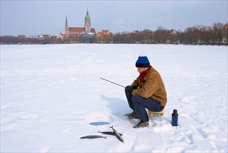 Man with winter cap or hat and warm clothes, sitting on a bucket, ice fishing on Theresienwiese at