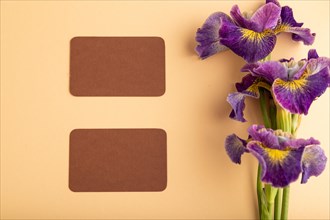 Brown business card with lilac iris flowers on orange pastel background. top view, flat lay, copy