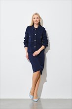 Elegant woman in navy blue button down jacket, skirt and blue heels posing against a white wall