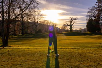 Female Golfer Teeing off with Her Golf Club Driver on Golf Course in Sunset and Lens Flare in