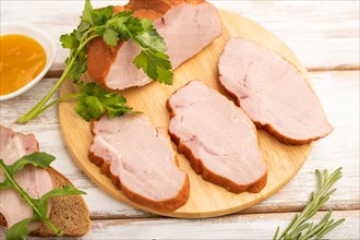 Smoked pork ham on cutting board with pepper and herbs on white wooden background. Side view, close